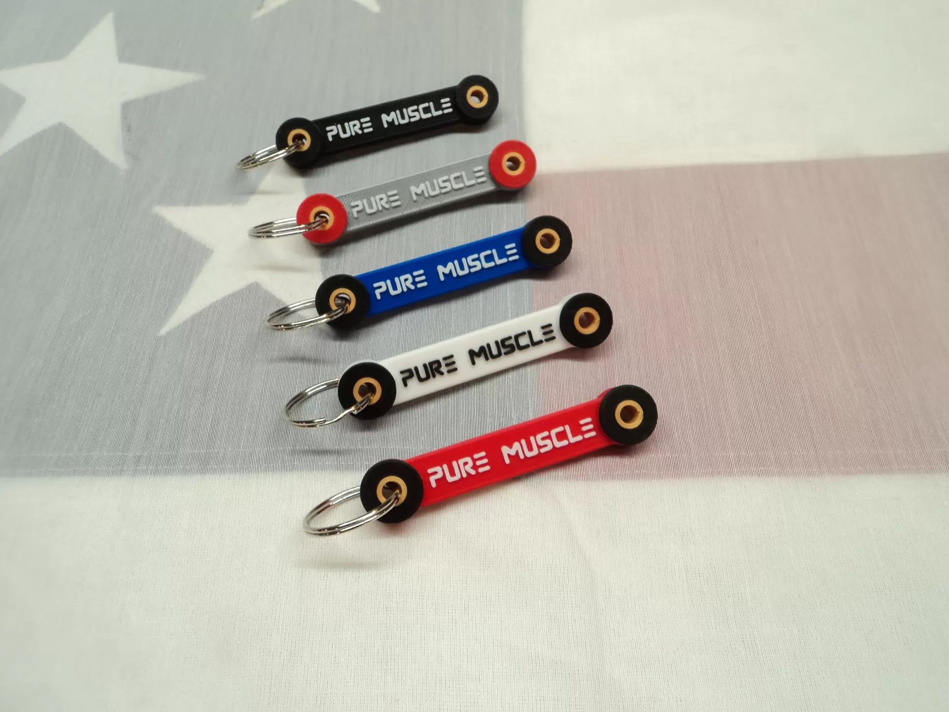 Five 'PURE MUSCLE' keychains in different colors arranged diagonally on a star-spangled background.