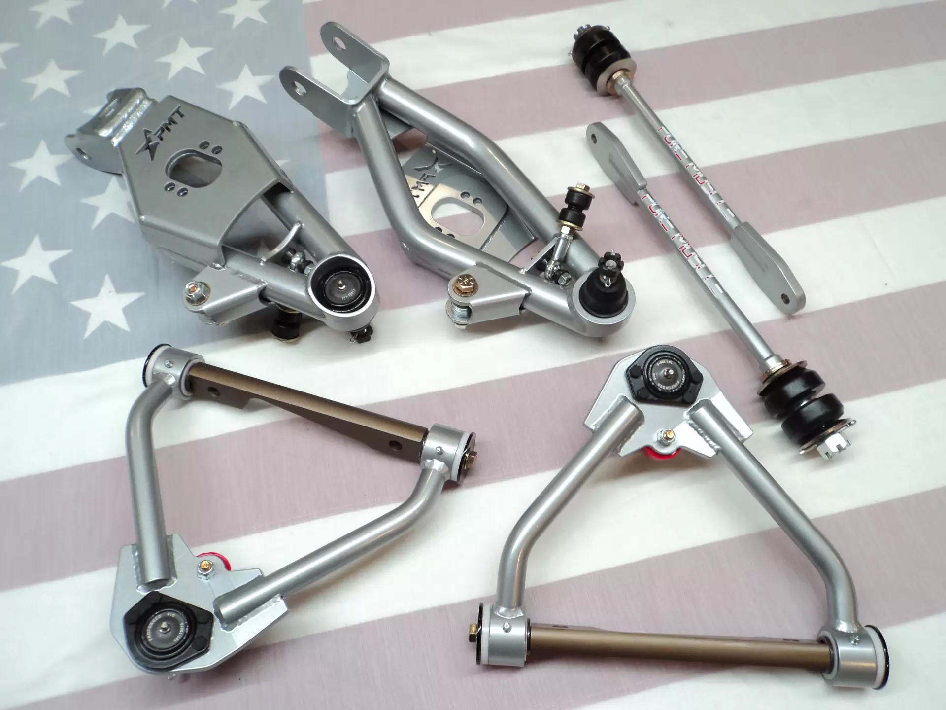 Suspension parts laid on a striped American flag background, including control arms, bushings, and brackets, ready for assembly.