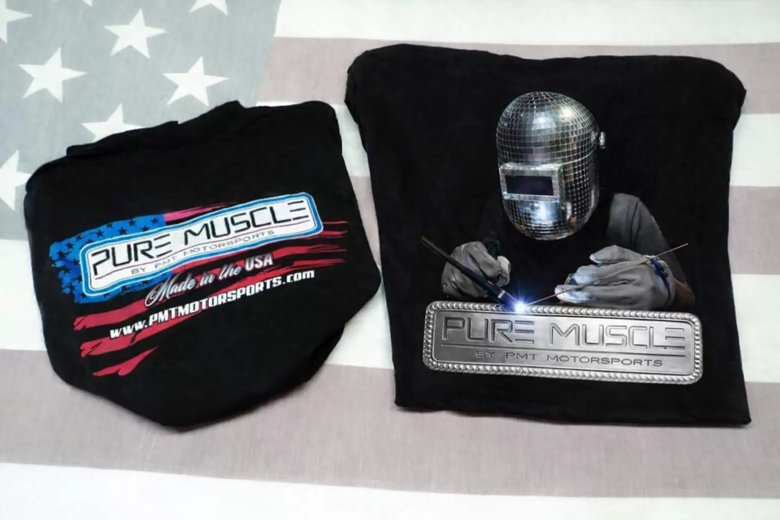 Two black shirts: left shows a blue-and-red Pure Muscle logo; right features a welding scene with a metallic badge reading 'Pure Muscle by PMT Motorsports'; background is a flag pattern.