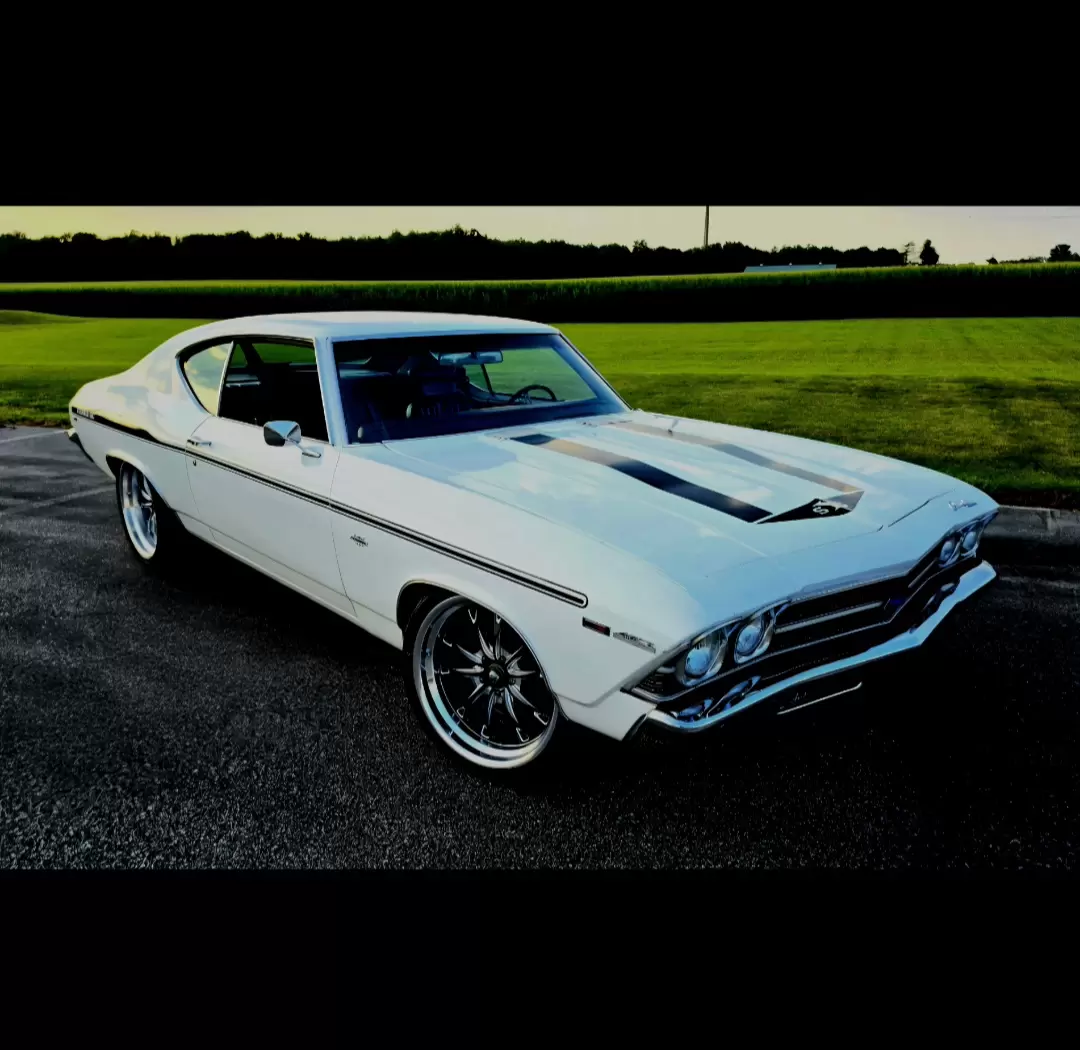 White classic American muscle car with a hood scoop and chrome trim parked on a paved lot, green fields in the background